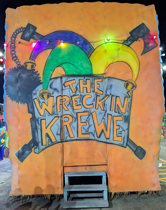 The back of the Wreckin' Krewe float — a jester hat above a banner reading 'The Wreckin' Krewe', with a wrecking ball on the left and an ax on the right.